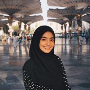 Portrait of a smiling young adult wearing a black hijab and polka dot dress under large, white umbrellas