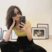 A fashionable, slender person in their early 20's sits in front of fashion photos and takes a selfie.