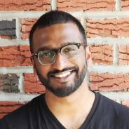 Portrait of a smiling person with a beard, glasses and closely cropped, dark hair wearing a black tshirt