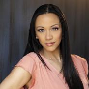 A Black woman of Caribbean descent with long, straight hair facing the camera. Leah wears a pink v-neck blouse and a thin silver necklace