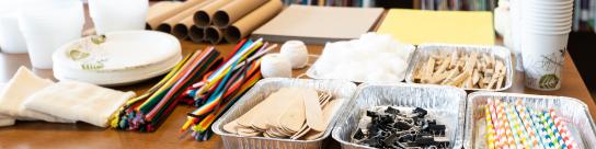 A library with a table full of classroom engineering design supplies: straws, paper, clips, pipecleaners, paper plates, tape and glue